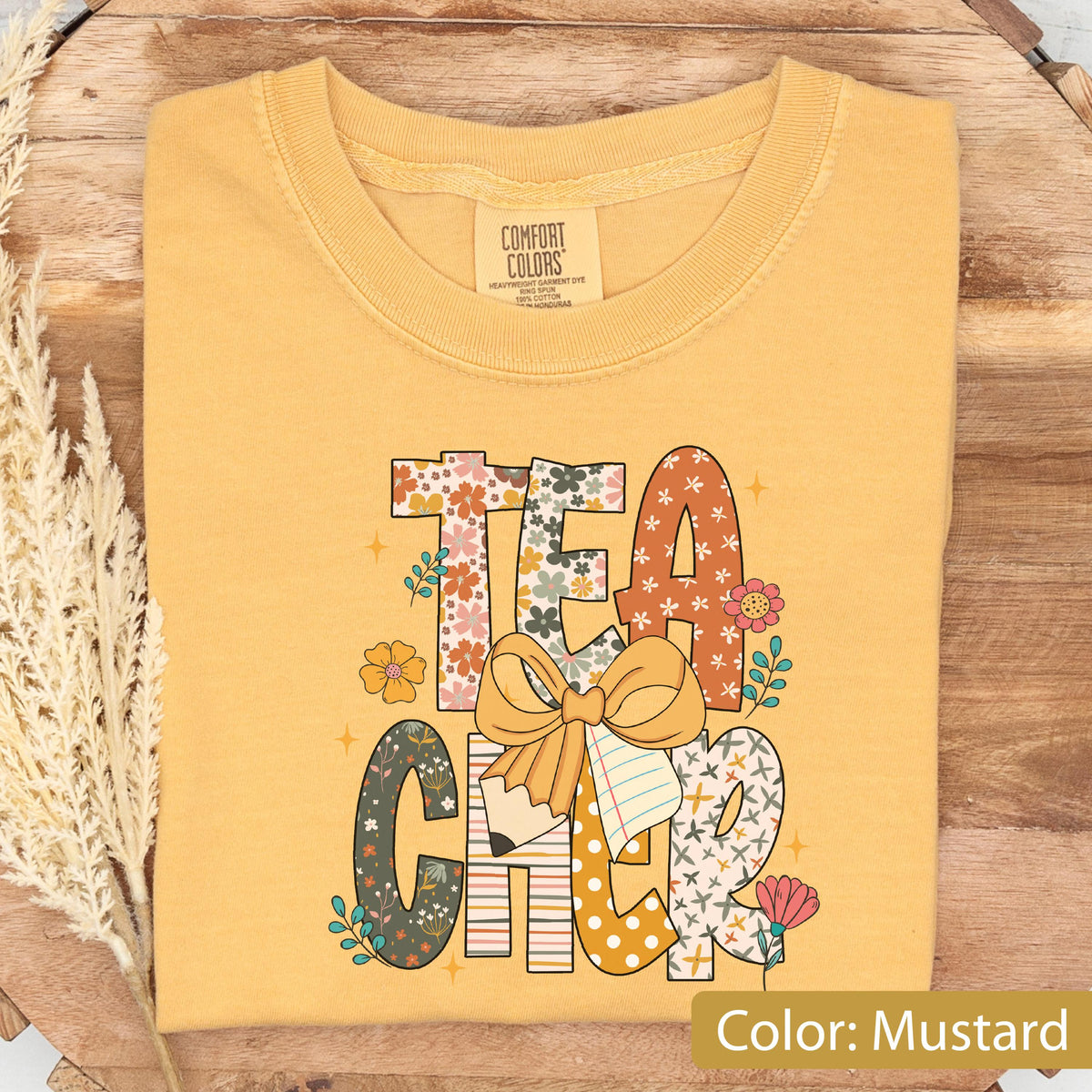 May include: Mustard-colored t-shirt with the word "TEACHER" in a floral and patterned design. The design includes a bow, pencil, and notebook. The shirt is folded on a wooden surface.