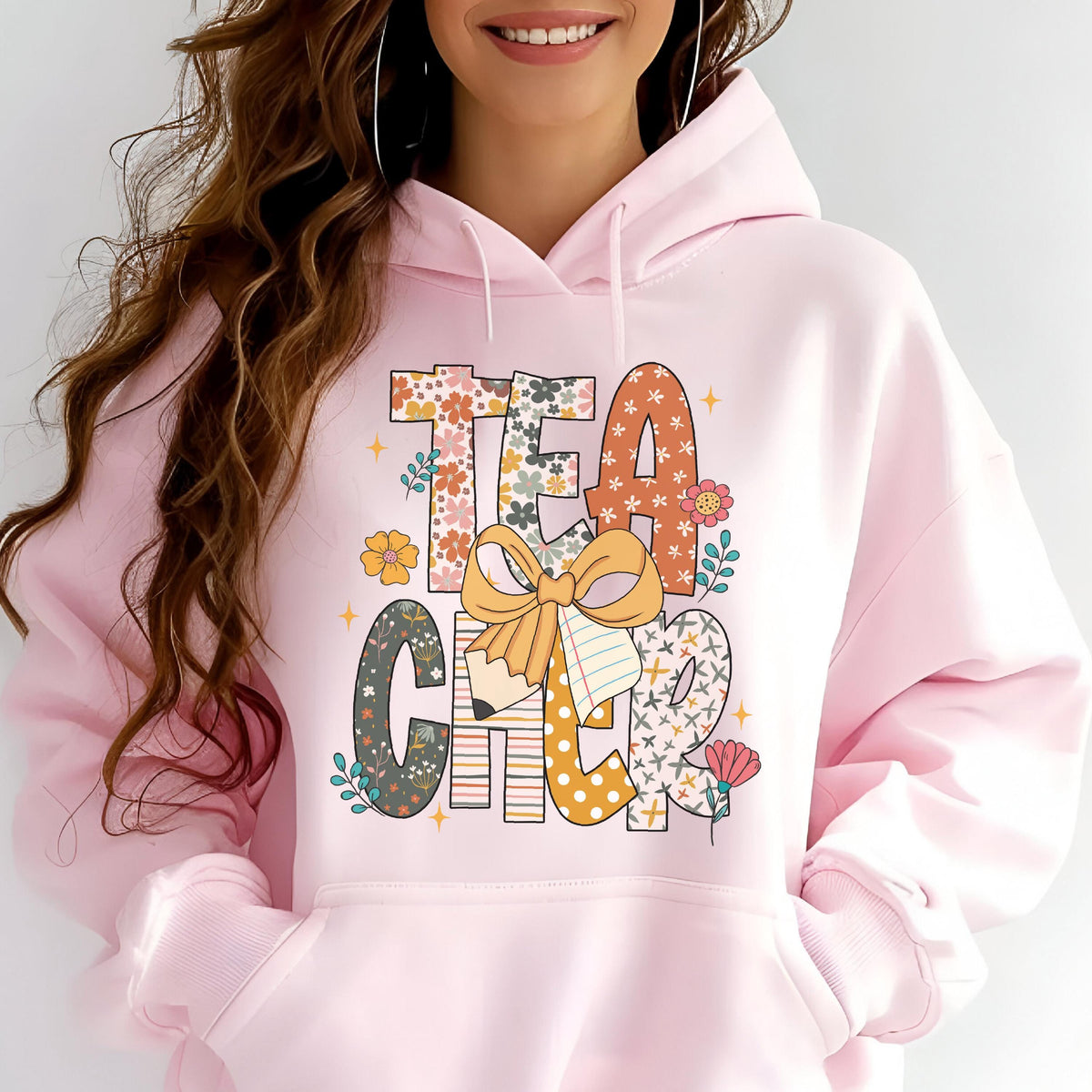 May include: A light pink hoodie with the word "TEACHER" in a colorful, floral design. The letters are decorated with flowers, a pencil, and a bow. The hoodie has a front pocket and a hood.