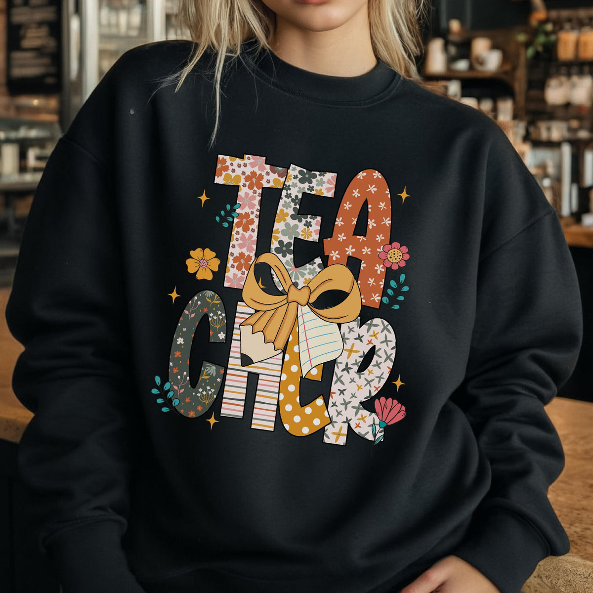 May include: Black sweatshirt with the word "TEACHER" in colorful, floral, and patterned letters. The letter "C" has a floral design, and the "H" is striped. A large bow is in the center of the word.