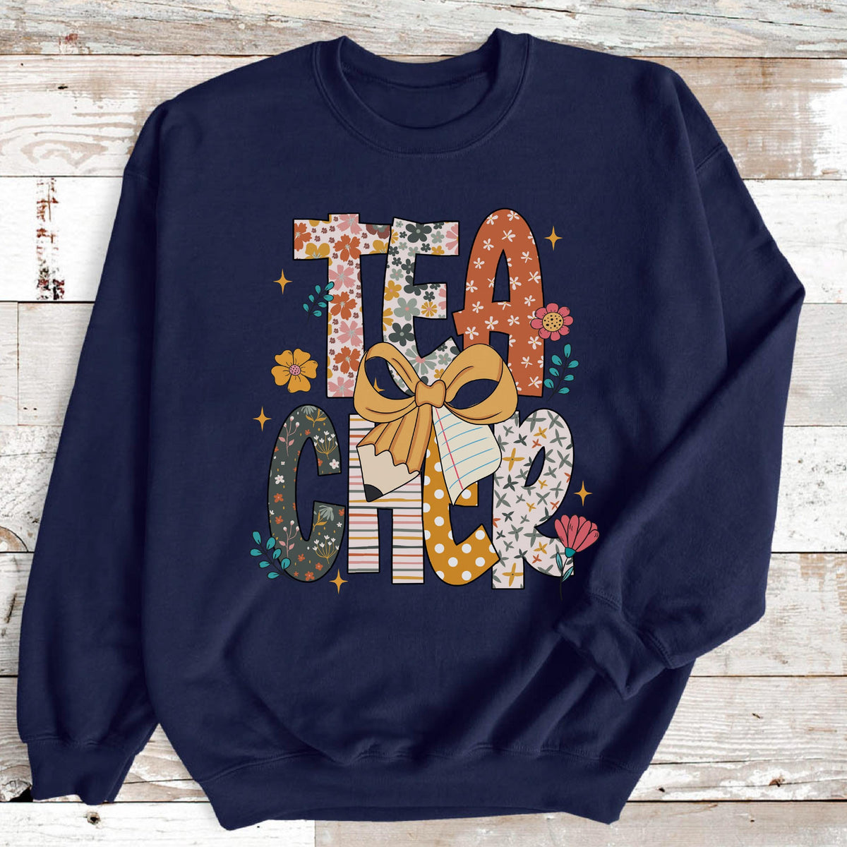 May include: Navy blue sweatshirt with the word "TEACHER" in a colorful, floral design. The letters are decorated with flowers, bows, and pencil graphics. A cute and stylish design for teachers.