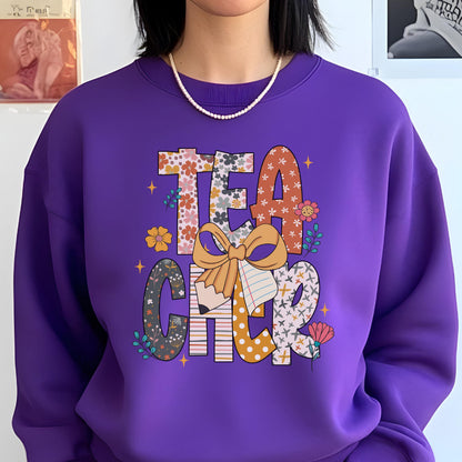 May include: A purple sweatshirt with the word "TEACHER" in a colorful, floral design. The letters are filled with various patterns, including flowers and polka dots. A large bow and pencil graphic are centered below the word.