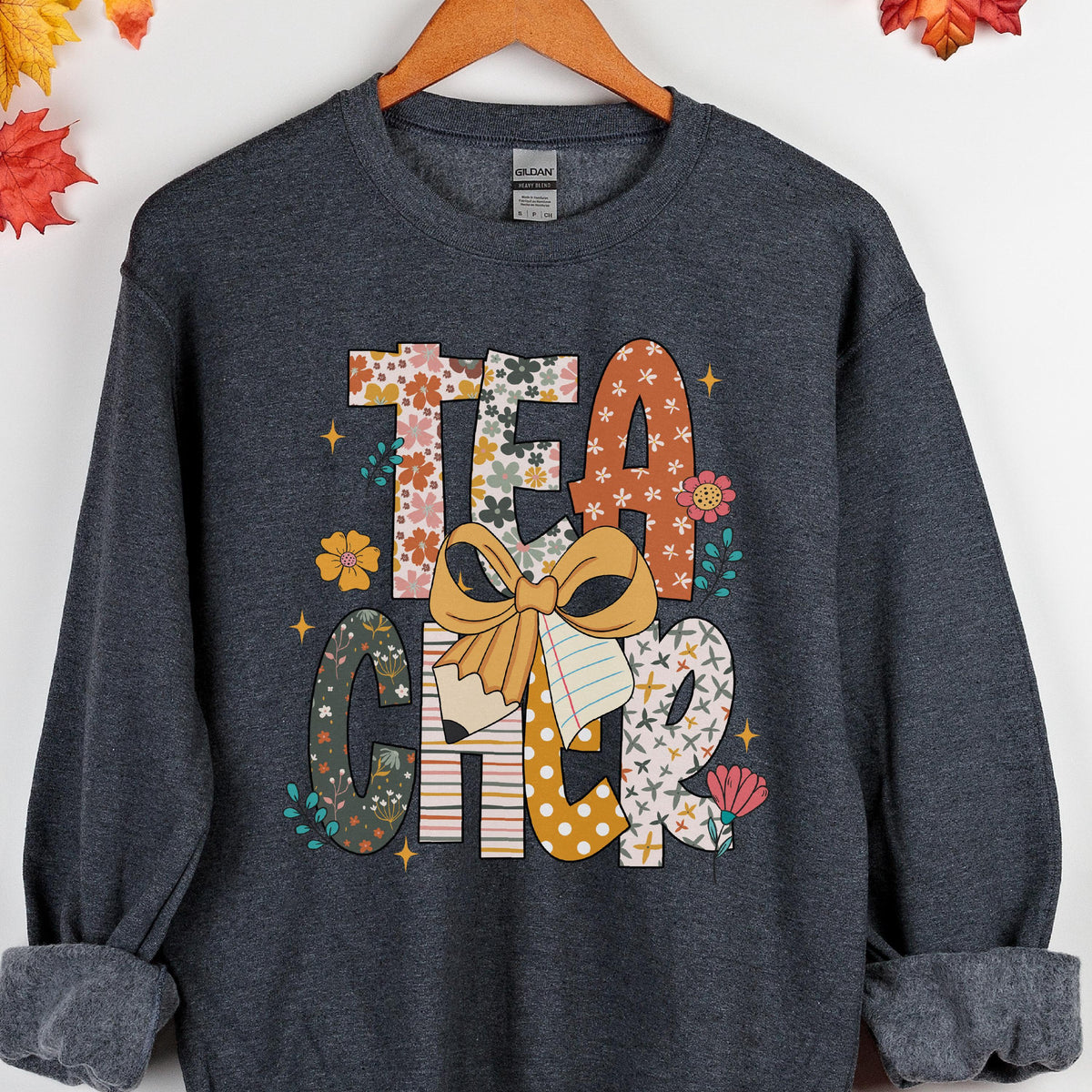 May include: Dark gray sweatshirt with the word "TEACHER" in a colorful floral design. The letters are decorated with flowers, bows, and patterns. The sweatshirt is hanging on a wooden hanger.