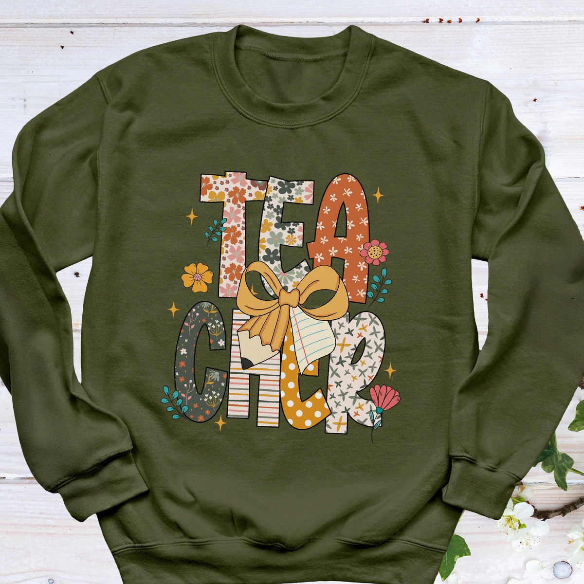 May include: An olive green sweatshirt with the word "TEACHER" in a colorful, floral, and patterned design. The letters are decorated with flowers, bows, and a pencil, creating a cheerful and educational theme.