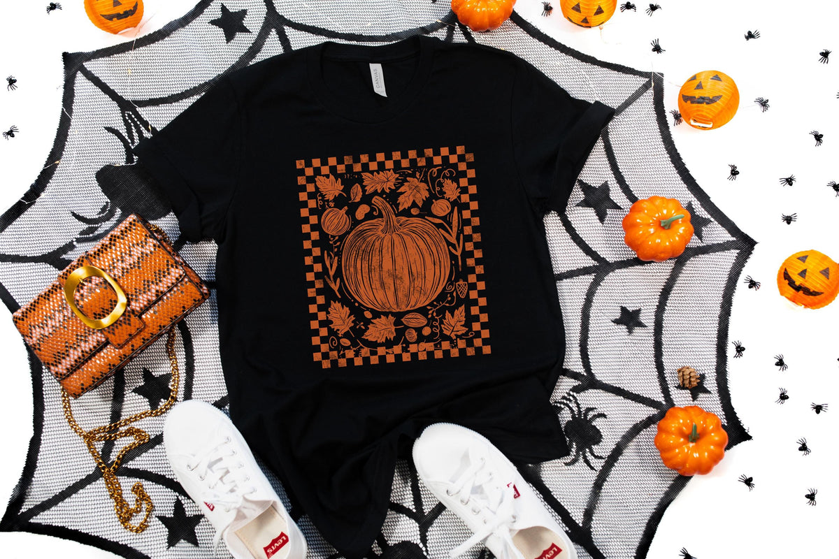 May include: Black t-shirt with an orange pumpkin graphic inside a checkered border. The image includes a matching purse, white sneakers, and Halloween decorations like pumpkins and a spiderweb.