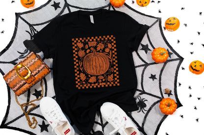 May include: Black t-shirt with an orange pumpkin graphic inside a checkered border. The image includes a matching purse, white sneakers, and Halloween decorations like pumpkins and a spiderweb.