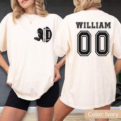 May include: Ivory-colored t-shirt with a black football graphic and the name "WILLIAM" on the front. The back of the shirt features the name "WILLIAM" above the number "00" in a black and white design. The shirt is a relaxed fit.
