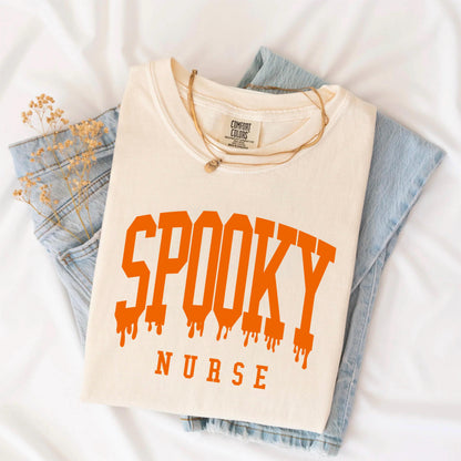 May include: A cream-colored short-sleeve t-shirt featuring the text SPOOKY in a large, orange, dripping varsity-style font, with the word NURSE printed in a smaller, plain orange font underneath. The shirt has a Comfort Colors brand label.