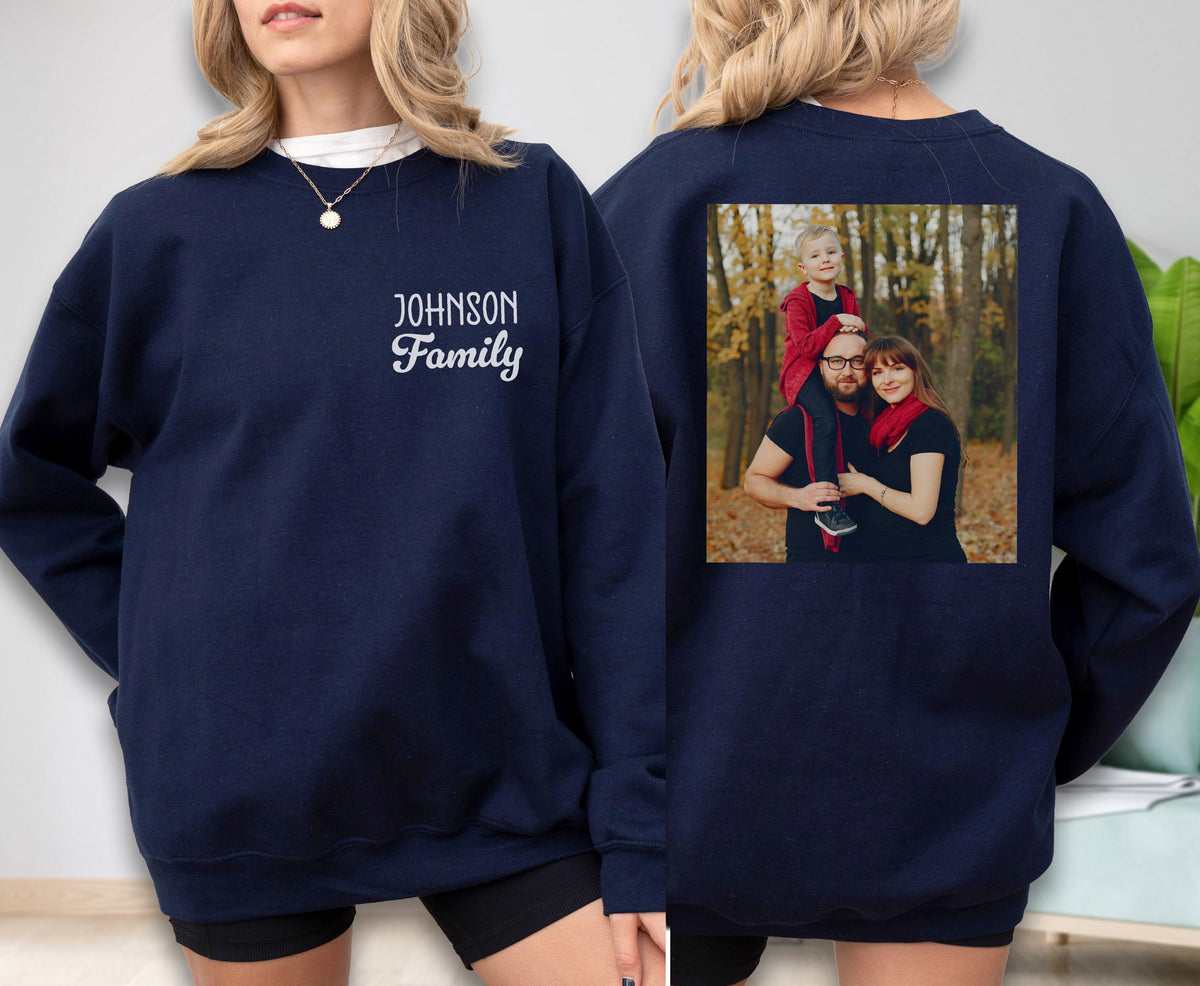 May include: Navy blue sweatshirt with the text "Johnson Family" in white script on the front. The back of the sweatshirt features a family photo. The photo shows a family of three in a forest setting.