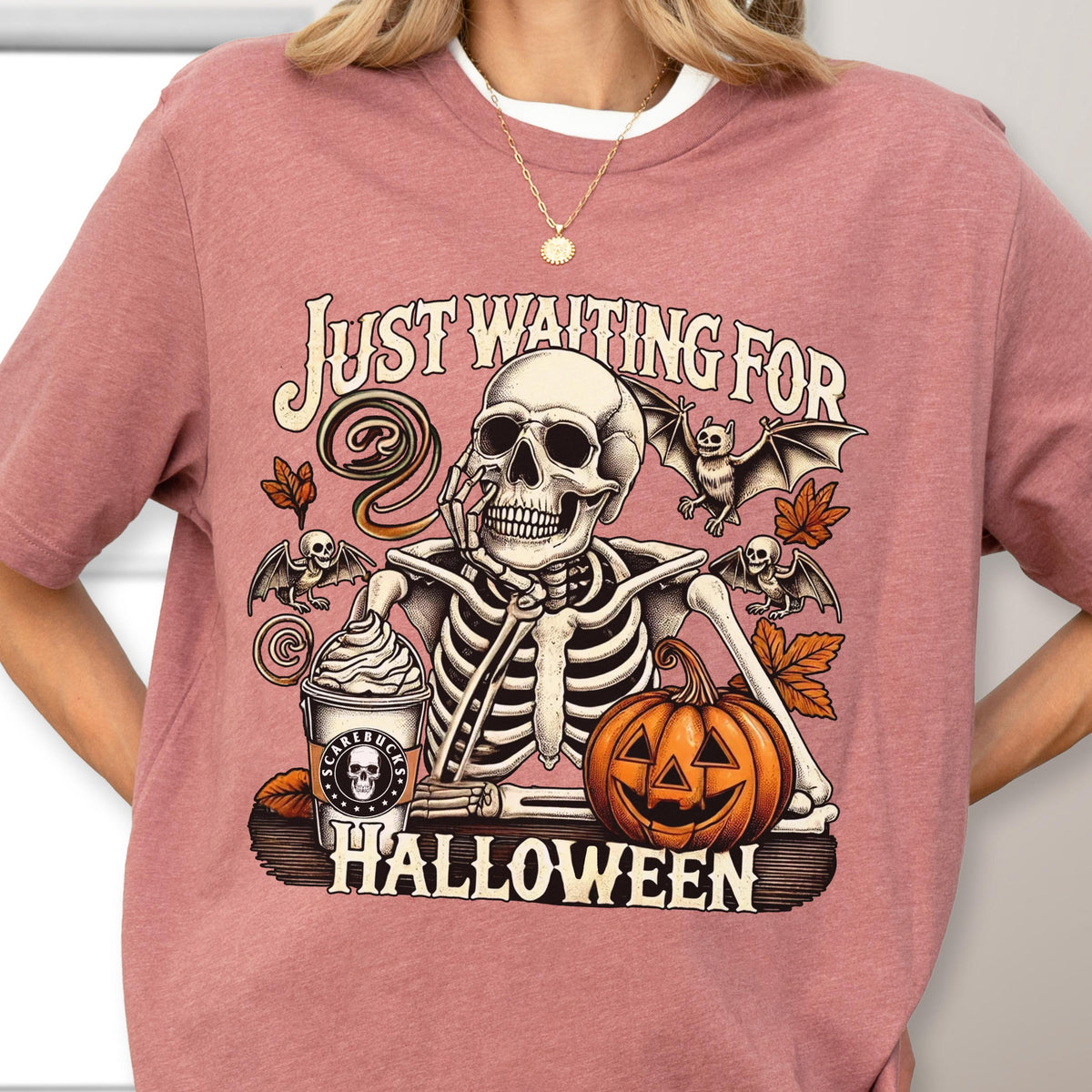 May include: Dusty rose t-shirt with a Halloween-themed graphic. The design features a skeleton with a coffee cup and a jack-o'-lantern, with the text "JUST WAITING FOR HALLOWEEN". Bats and autumn leaves complete the design.