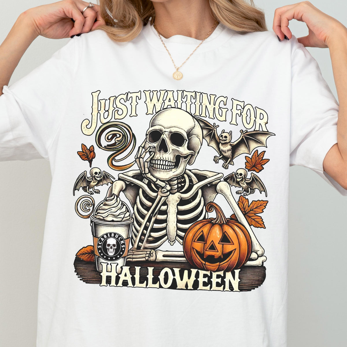 May include: White t-shirt with a Halloween-themed graphic. The design features a skeleton with a coffee cup, pumpkin, and bats, with the text "Just Waiting For Halloween". The graphic is in shades of orange, brown, and black.