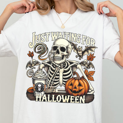 May include: White t-shirt with a Halloween-themed graphic. The design features a skeleton with a coffee cup, pumpkin, and bats, with the text "Just Waiting For Halloween". The graphic is in shades of orange, brown, and black.
