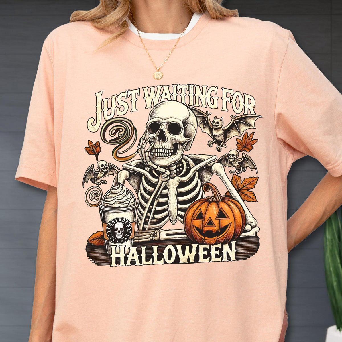 May include: A light pink t-shirt with a Halloween-themed graphic. The graphic features a skeleton, bats, a pumpkin, and a coffee cup. The text "JUST WAITING FOR HALLOWEEN" is printed above and below the skeleton.