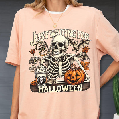 May include: A light pink t-shirt with a Halloween-themed graphic. The graphic features a skeleton, bats, a pumpkin, and a coffee cup. The text "JUST WAITING FOR HALLOWEEN" is printed above and below the skeleton.