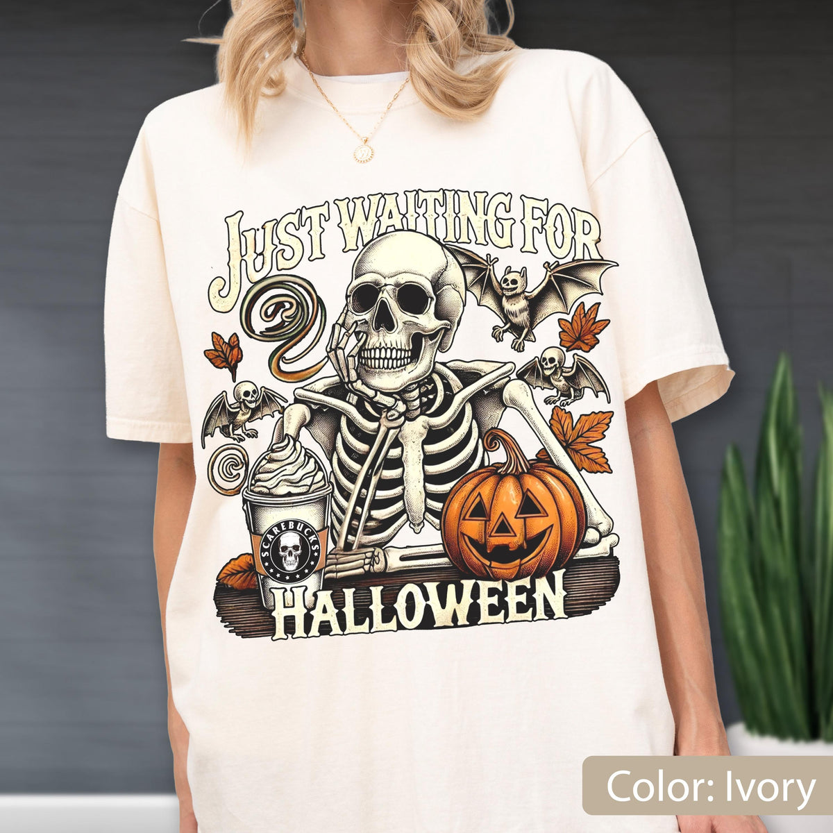 May include: An ivory t-shirt featuring a graphic of a skeleton resting its chin on its hand next to a pumpkin and a coffee cup. The text reads "JUST WAITING FOR HALLOWEEN" and "SCARIBUCKS" on the cup. The design includes bats and autumn leaves.