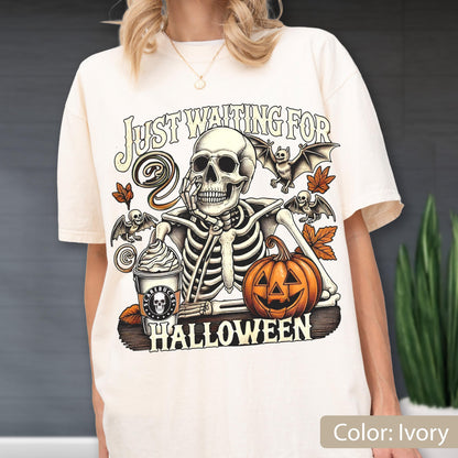 May include: An ivory t-shirt featuring a graphic of a skeleton resting its chin on its hand next to a pumpkin and a coffee cup. The text reads "JUST WAITING FOR HALLOWEEN" and "SCARIBUCKS" on the cup. The design includes bats and autumn leaves.