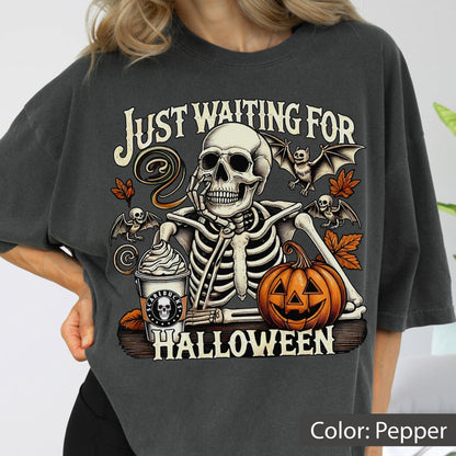 May include: A pepper-colored t-shirt featuring a graphic of a skeleton resting its chin on its hand next to a pumpkin and a coffee cup. The text reads "JUST WAITING FOR HALLOWEEN" with illustrations of bats and autumn leaves surrounding the central skeleton figure.