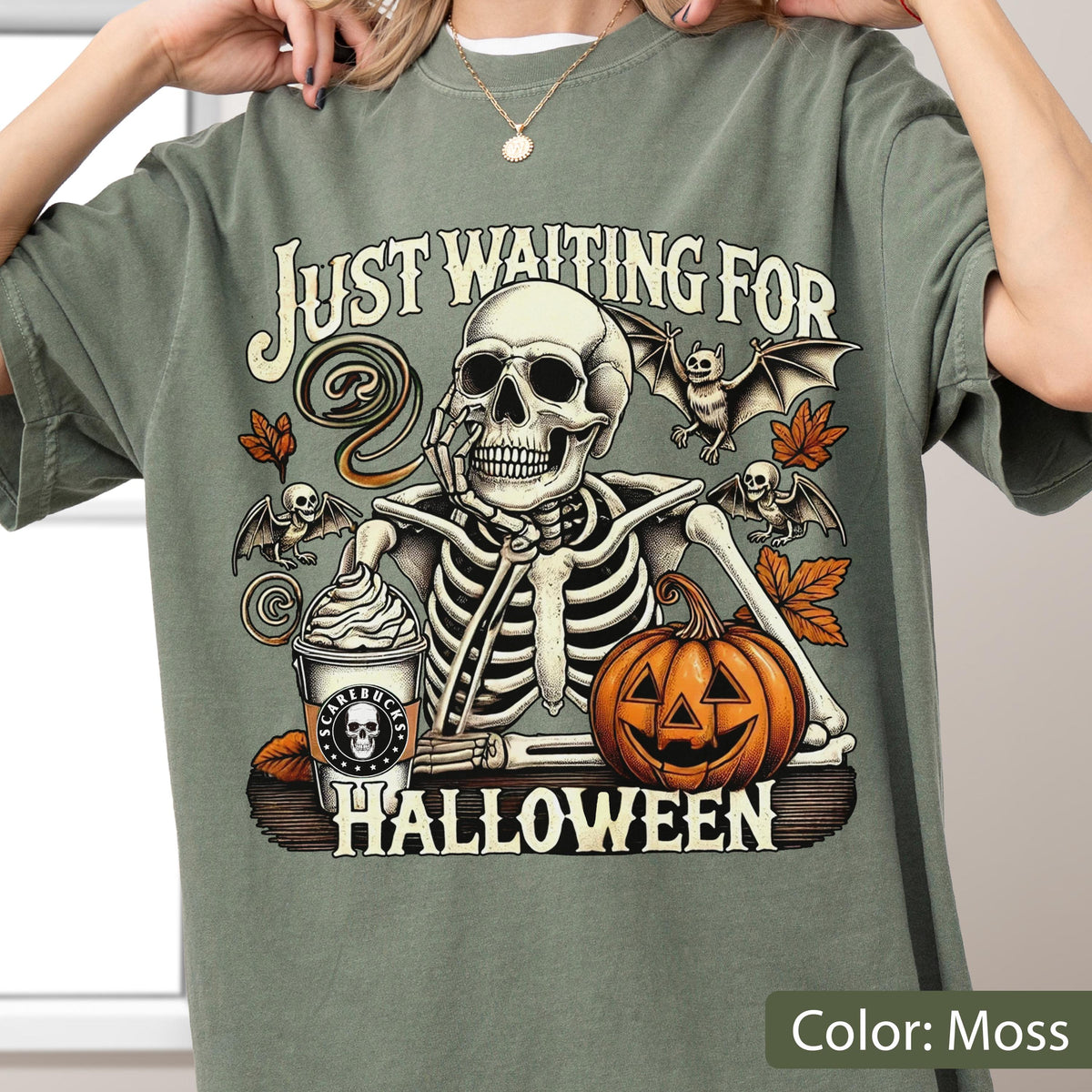 May include: A moss green t-shirt featuring a graphic of a skeleton resting its chin on its hand. The design includes a coffee cup labeled SCAREBUCKS, a jack-o'-lantern, bats, and autumn leaves, with the text JUST WAITING FOR HALLOWEEN printed.