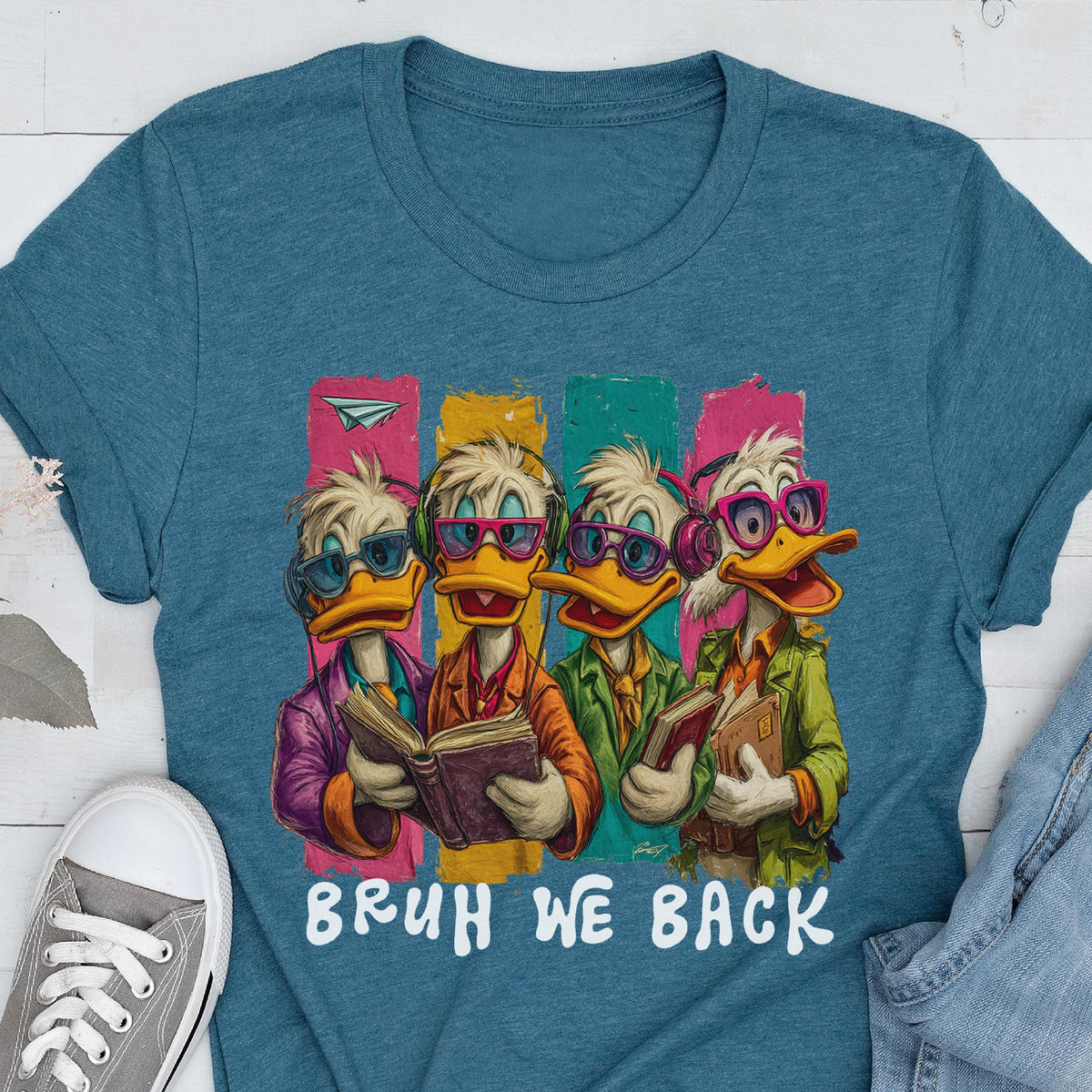 May include: Teal t-shirt featuring a cartoon illustration of four ducks wearing glasses and headphones, each holding a book. The ducks stand in front of colorful vertical stripes. The text "BRUH WE BACK" is printed below the illustration.