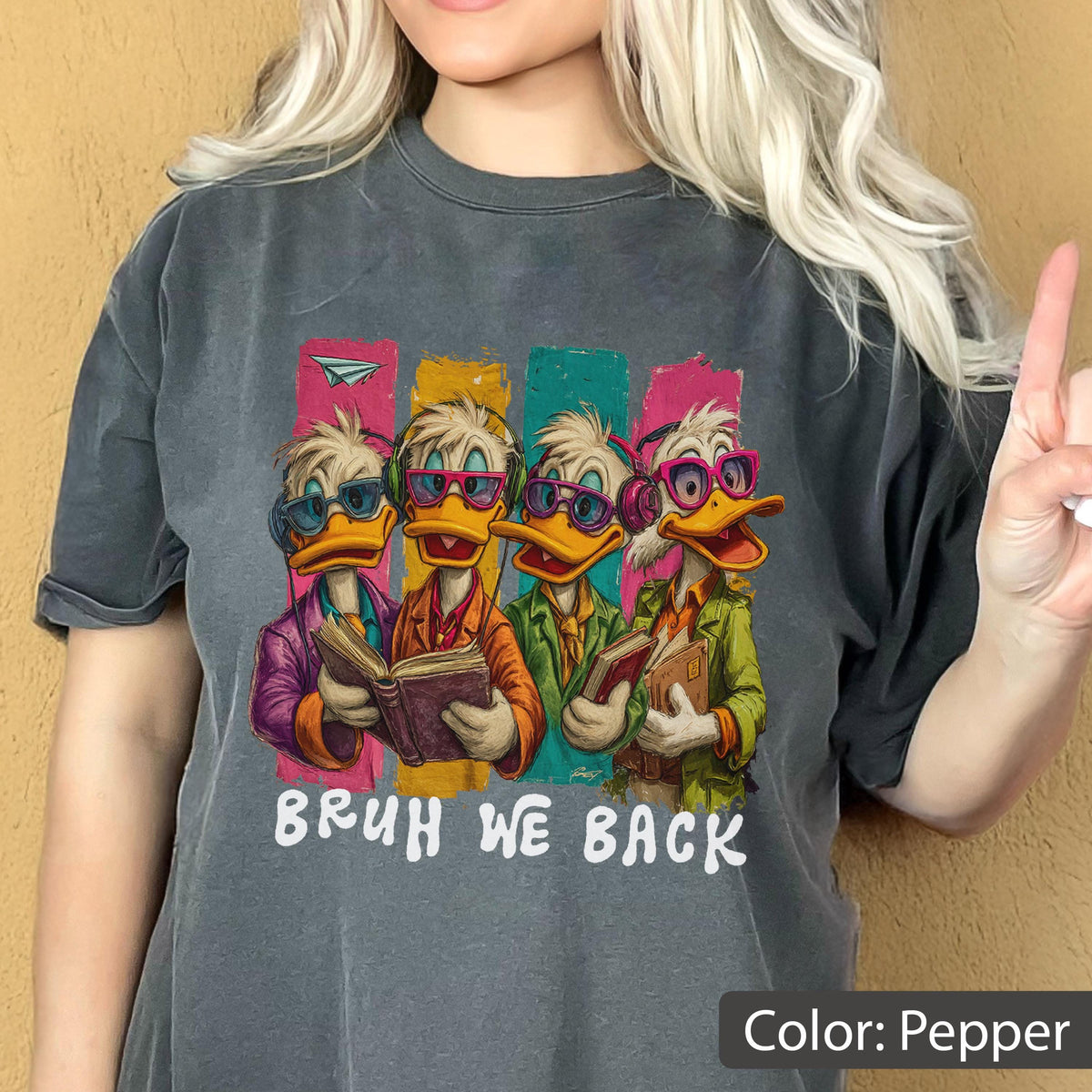 May include: Pepper-colored t-shirt featuring a cartoon illustration of four ducks wearing glasses and reading books. The ducks are set against colorful vertical stripes. The text "BRUH WE BACK" is printed below the ducks.