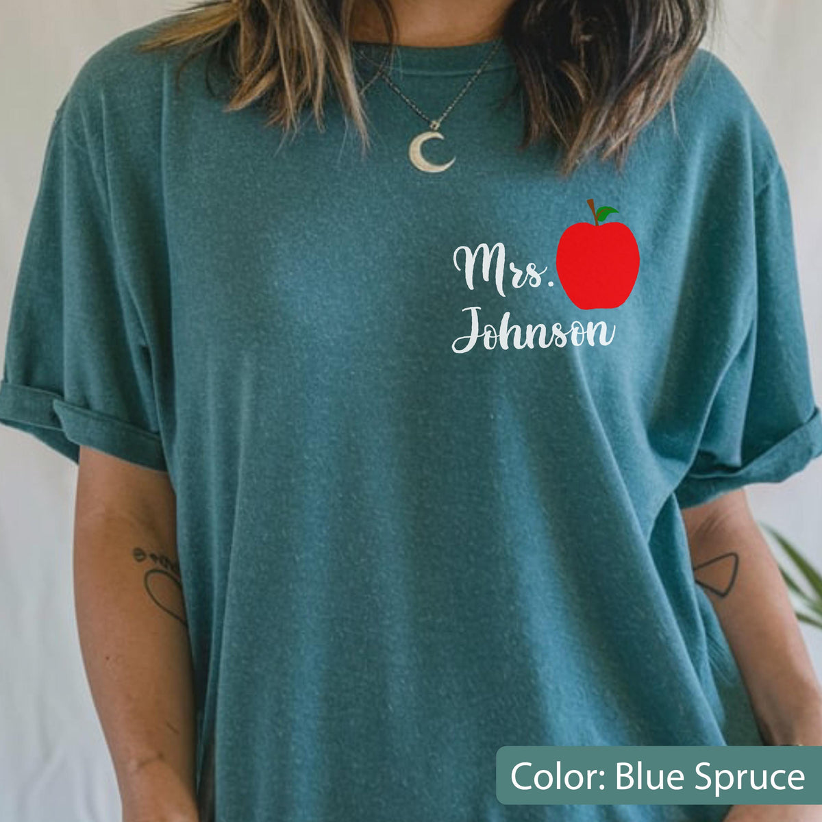 May include: A blue spruce short-sleeve t-shirt featuring a graphic on the left chest area. The graphic consists of a bright red apple with a green leaf and the white cursive text that reads Mrs. Johnson positioned directly below the apple.
