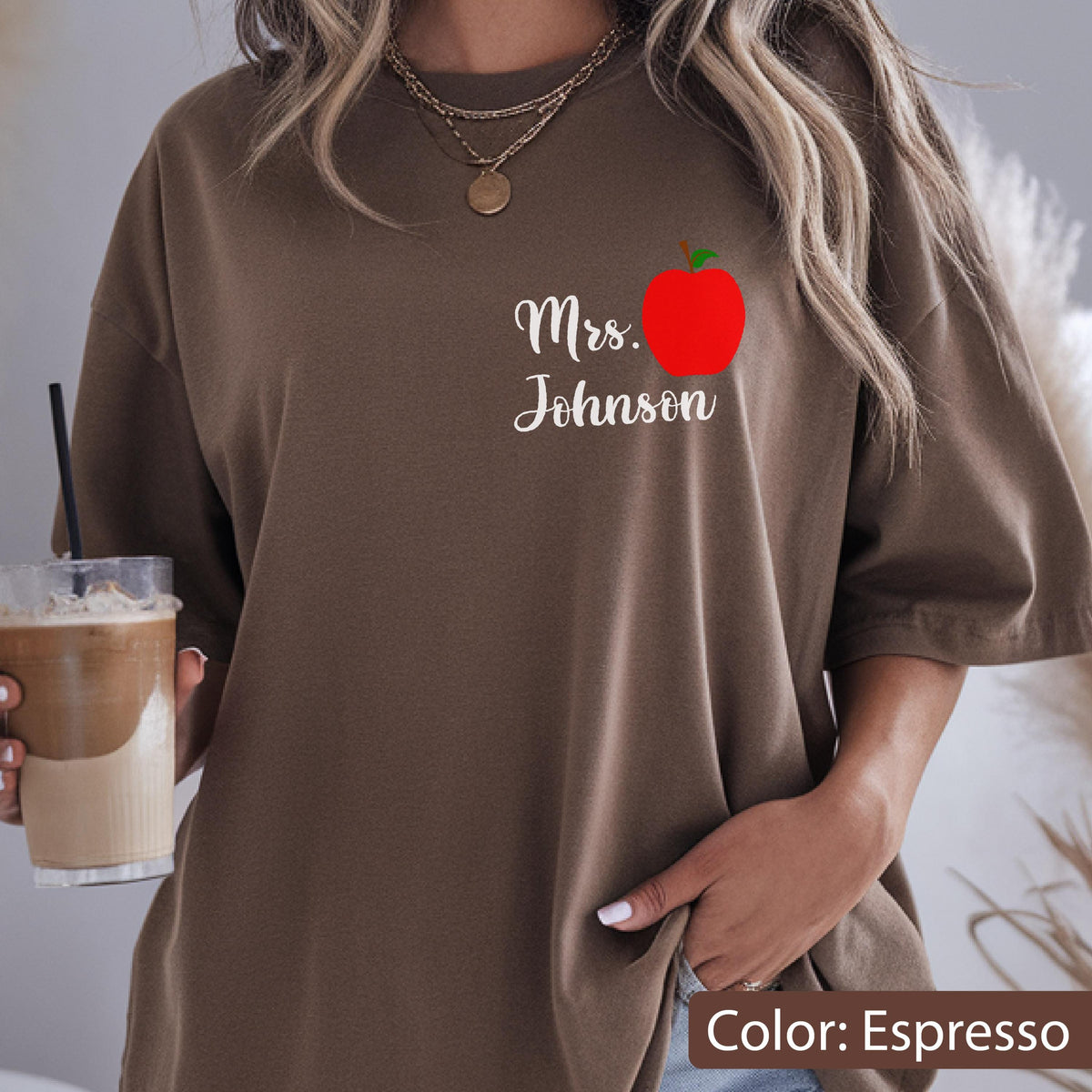 May include: An espresso-colored t-shirt featuring a graphic on the left chest area. The graphic includes a bright red apple illustration positioned above the white cursive text that reads Mrs. Johnson. The shirt has short sleeves and a crew neckline.