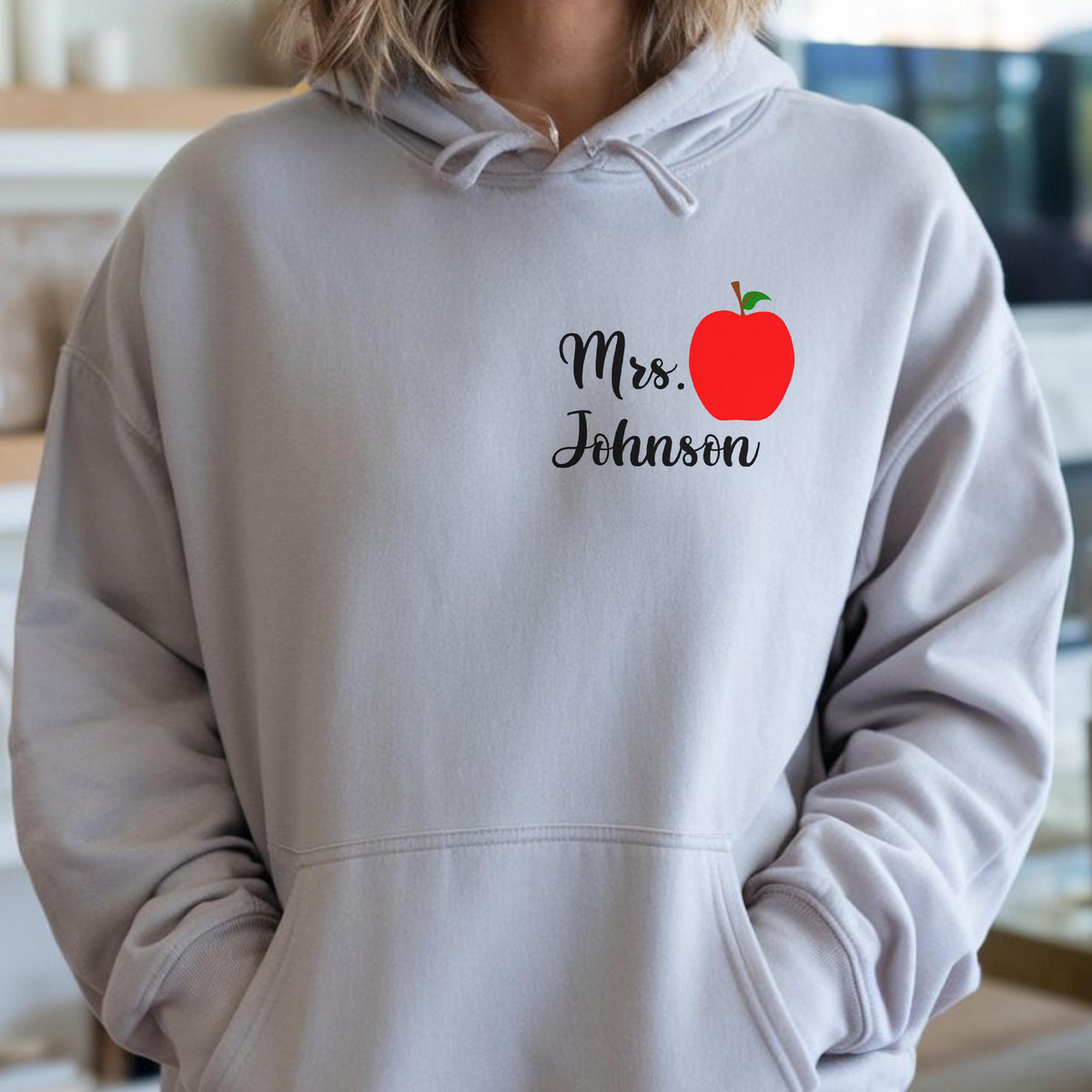 May include: A light gray hoodie with a front pocket and drawstring hood. The hoodie features the text "Mrs. Johnson" in black script, accompanied by a red apple graphic with a green leaf.