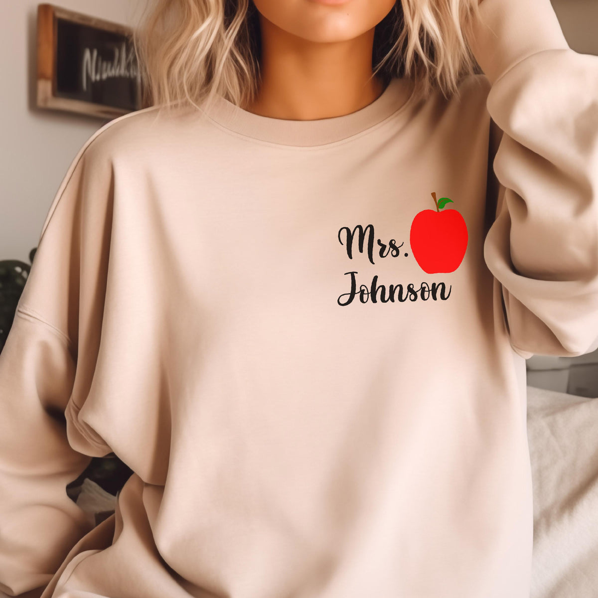May include: Beige sweatshirt with the text "Mrs. Johnson" in black script, accompanied by a red apple graphic with a green leaf. The sweatshirt is a neutral color and has a crew neck.