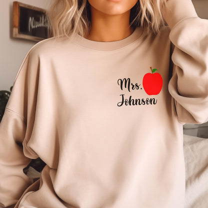 May include: Beige sweatshirt with the text "Mrs. Johnson" in black script, accompanied by a red apple graphic with a green leaf. The sweatshirt is a neutral color and has a crew neck.