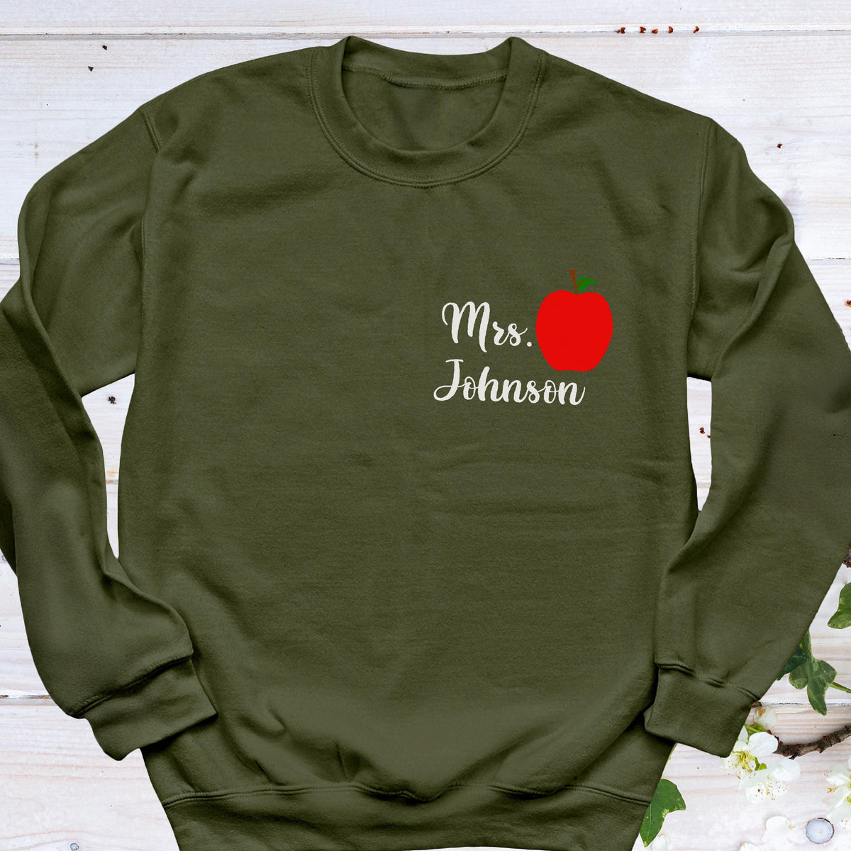 May include: Olive green sweatshirt with the text "Mrs. Johnson" in white script, accompanied by a red apple graphic. The sweatshirt is a crew neck style.