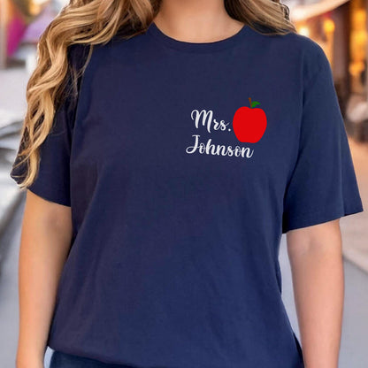May include: Navy blue t-shirt with the text "Mrs. Johnson" in a cursive font, accompanied by a red apple graphic with a green stem. The shirt is a solid color and has short sleeves.