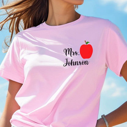 May include: Pink t-shirt with the text "Mrs. Johnson" in black script and a red apple graphic. The shirt is a light pink color and has short sleeves. The background is a blurred blue sky.