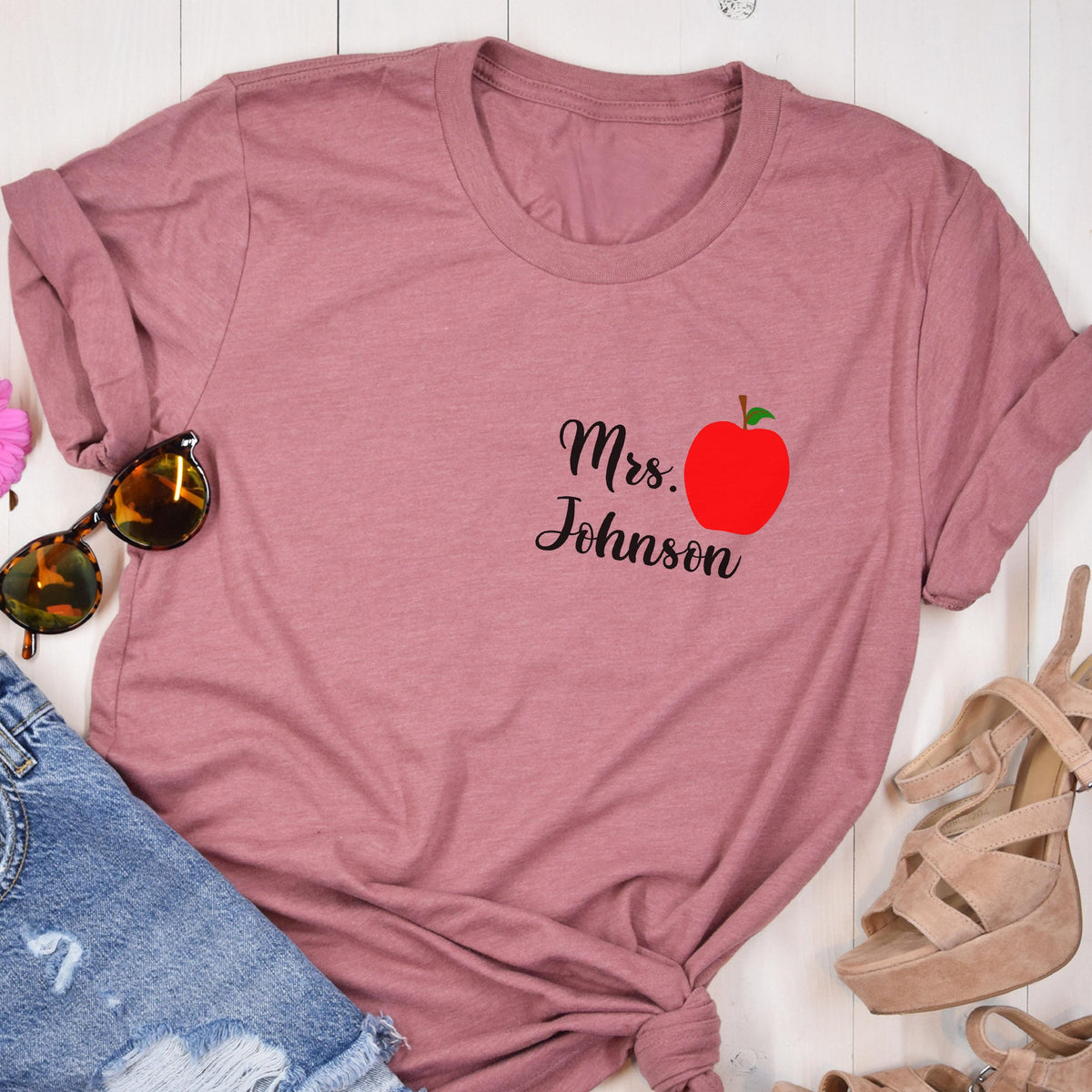 May include: Dusty rose t-shirt with rolled sleeves. Features the text "Mrs. Johnson" in black script, accompanied by a red apple graphic. Sunglasses and denim shorts are visible in the background.