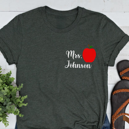 May include: Dark gray t-shirt with the text "Mrs. Johnson" in white script, accompanied by a red apple graphic. The shirt has a crew neck and short sleeves, suitable for casual wear.