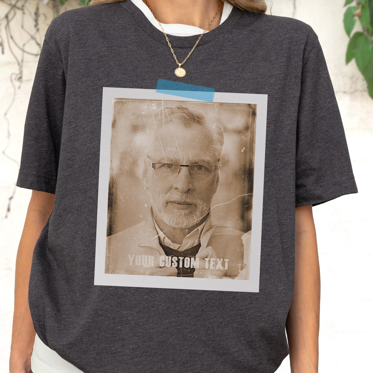 May include: Dark gray t-shirt with a sepia-toned photo print of a man wearing glasses. The photo is taped to the shirt with blue tape and has the text "YOUR CUSTOM TEXT" at the bottom. The shirt has a white crew neck collar.