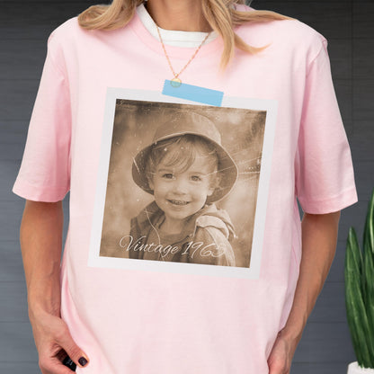 May include: A light pink t-shirt with a vintage sepia-toned photograph of a child wearing a hat. The photograph is taped to the shirt with a blue piece of tape. The text "Vintage 1965" is visible on the photograph.
