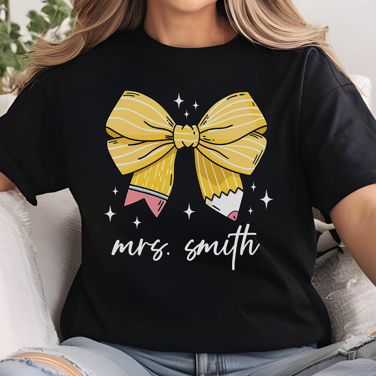 May include: Black t-shirt featuring a yellow bow made of pencils, with the text "mrs. smith" in white script. The bow design includes pink and white accents, and is surrounded by small white stars. A casual, everyday tee.