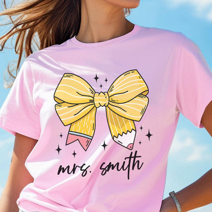 May include: Pink t-shirt featuring a large yellow bow and pencil design. The bow is detailed with white lines, and the pencils have pink erasers. The text "mrs. smith" is written in black script below the bow, with star accents.