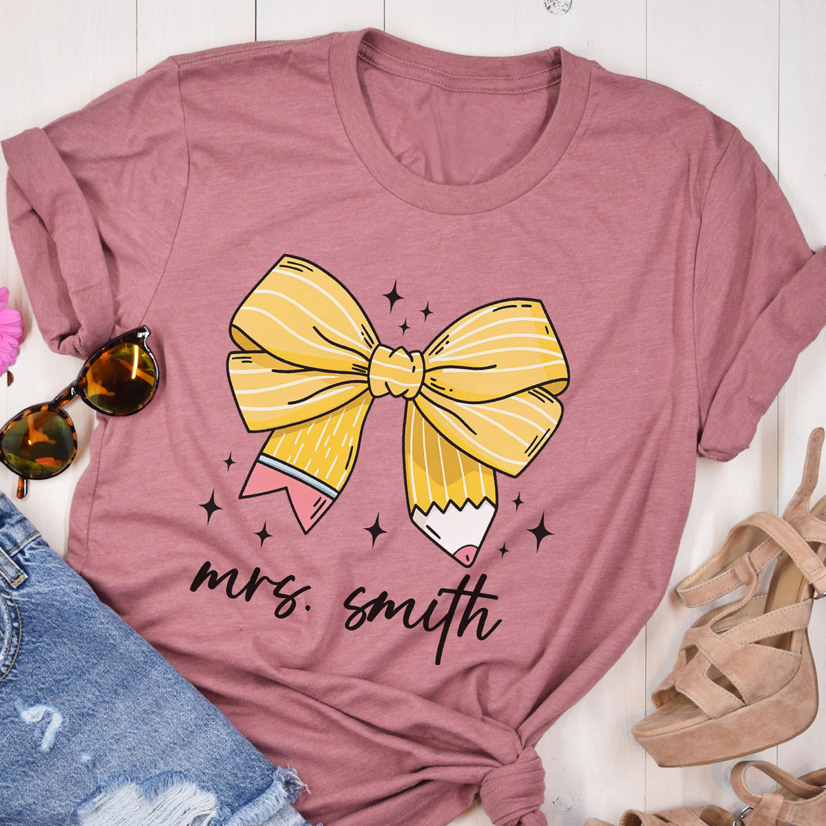 May include: A dusty rose t-shirt featuring a yellow bow made of pencils with the text "mrs. smith" in black script. The bow design includes white stripes and pink and blue accents. The shirt is styled with sunglasses and denim shorts.