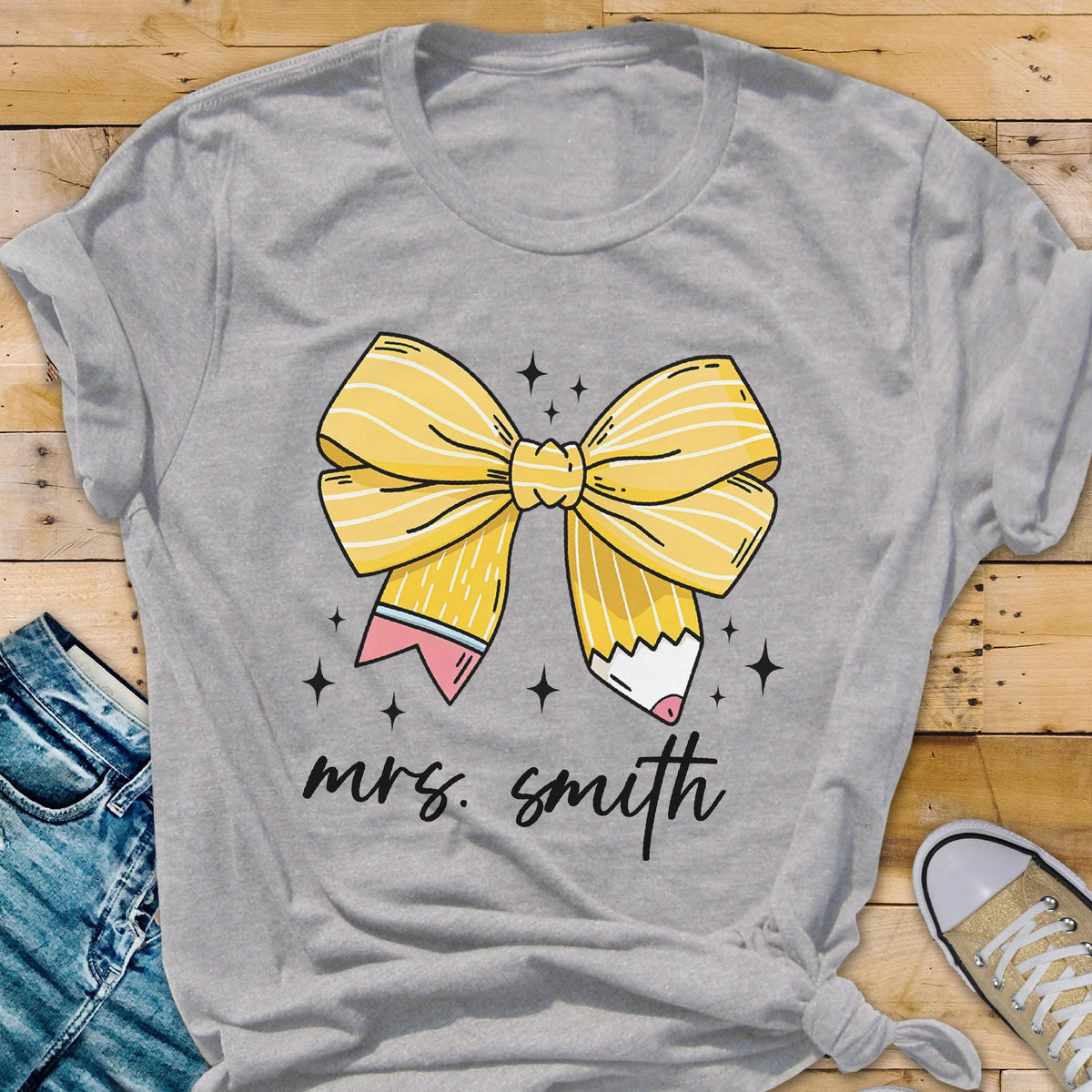 May include: A gray t-shirt featuring a yellow bow design made of pencils. The pencils have pink and white accents. The text "mrs. smith" is written in black script below the bow.