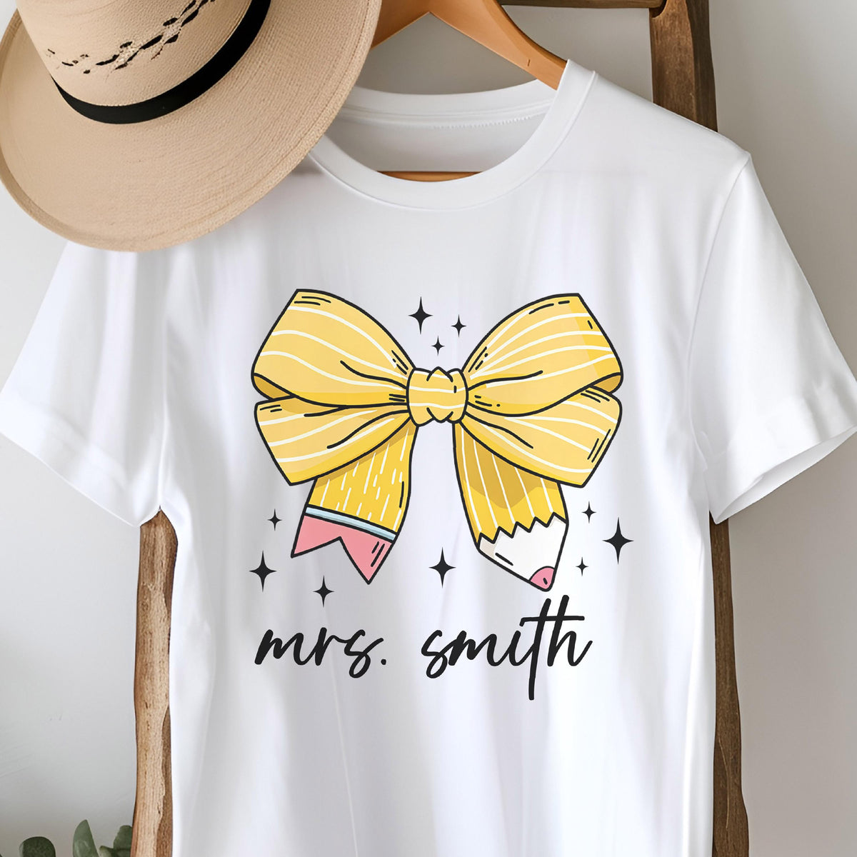 May include: White t-shirt featuring a large yellow bow design with pencil details and the text "mrs. smith" in black script. The bow has yellow stripes and is accented with small star shapes. A tan hat hangs on a wooden hanger.