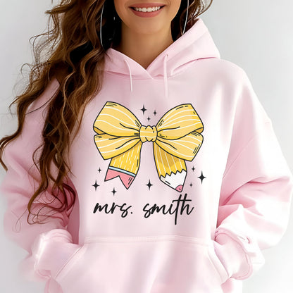 May include: A light pink hoodie featuring a large yellow bow with pencil details and the text "mrs. smith" in black script. The bow has a pencil design on each end, with pink and white accents. Small black stars are scattered around the bow.