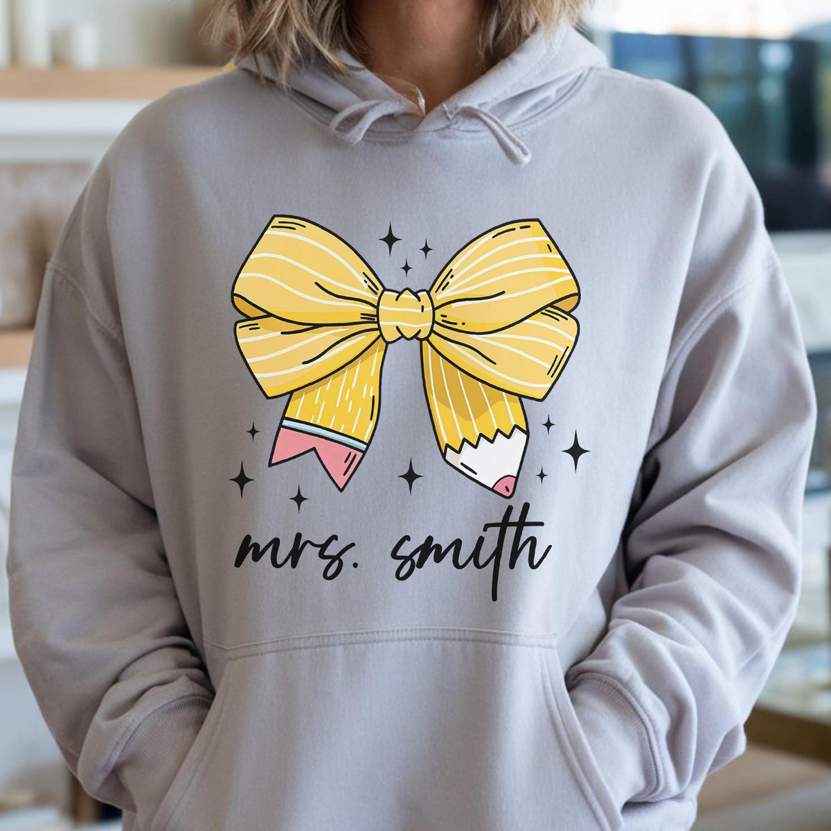 May include: A light gray hoodie featuring a large yellow bow with pencil details and the text "mrs. smith" in black script. The bow has white stripes and is accented with small black star shapes.