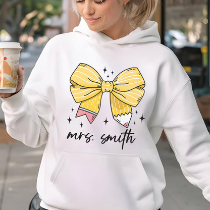 May include: White hoodie featuring a large yellow bow design with a pencil on each side. The text "mrs. smith" is written in a cursive font below the bow. The design is accented with small black star shapes.