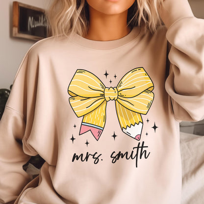 May include: Beige sweatshirt featuring a large yellow bow with pencil accents and the text "mrs. smith" in black script. The bow design includes a pencil with a pink eraser and blue stripes. Small black stars surround the bow.