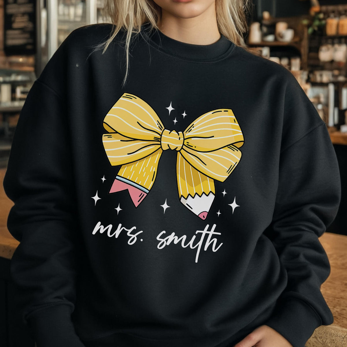 May include: Black sweatshirt featuring a large yellow bow design with pencil accents and the text "mrs. smith" in white script. The bow has yellow and white stripes. The design includes small white star accents.