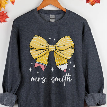 May include: A dark gray sweatshirt featuring a large yellow bow and pencil design. The bow is centered, with a pencil on each side. The text "mrs. smith" is written in cursive below the bow. White star accents are scattered around the bow.