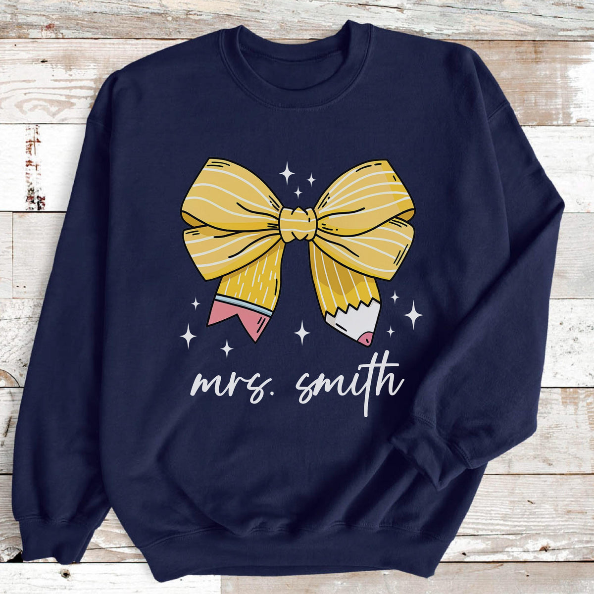 May include: Navy blue sweatshirt featuring a large yellow bow design made to look like a pencil. The bow is centered on the shirt, with the text "mrs. smith" in white script below. White star accents are scattered around the bow.