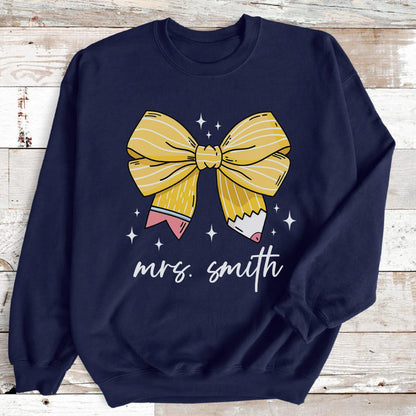 May include: Navy blue sweatshirt featuring a large yellow bow design made to look like a pencil. The bow is centered on the shirt, with the text "mrs. smith" in white script below. White star accents are scattered around the bow.