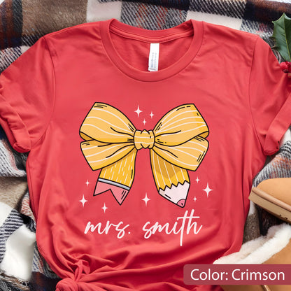 May include: Crimson-colored t-shirt featuring a yellow pencil bow design with the text "mrs. smith" in white. The bow has pencil-shaped ends with pink erasers. White star accents surround the bow.