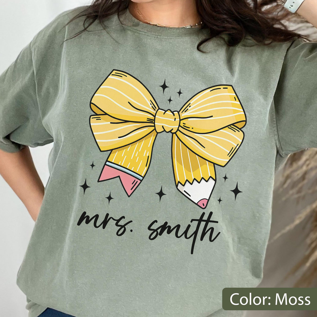 May include: Moss green t-shirt featuring a large yellow bow made of pencils. The pencils have pink erasers and are decorated with black stars. The name "mrs. smith" is written in black script below the bow.